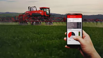ao fundo, pulverizador Kuhn em campo trabalhando e à frente, mão com celular na mão e sistema de telemetria da máquina aberto mostrando dados