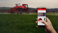 ao fundo, pulverizador Kuhn em campo trabalhando e à frente, mão com celular na mão e sistema de telemetria da máquina aberto mostrando dados